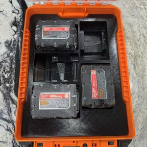 May include: An open orange plastic storage case with a black interior insert containing two black Milwaukee 6.0 Ah battery packs. The case features a clear hinged lid and molded compartments designed to hold the batteries securely in place.