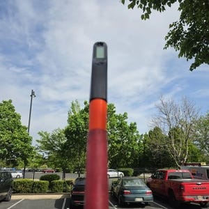 May include: A red and orange pole with a black top and a small digital display. The pole is held up against a blue sky with some trees and a parking lot in the background.