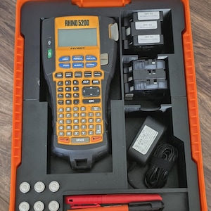 May include: An orange and black Rhino 5200 label maker kit. The kit includes the label maker, label cartridges, a power adapter, and pens. The label maker has an orange keypad and a black display screen. The kit is stored in an orange case.