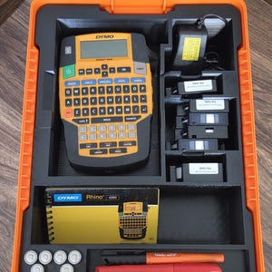 May include: An orange and black DYMO Rhino 4200 label maker kit. The kit includes the label maker, label cartridges, a power adapter, markers, and a user manual, all stored in an orange case.
