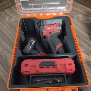 May include: An open orange and black tool case containing a red and black drill, a battery, and a red tool box. The case has a clear top and is sitting on a wooden surface.