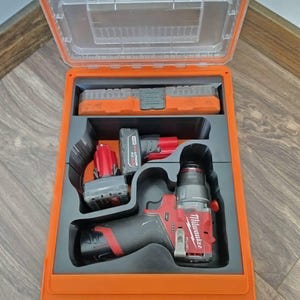 May include: An orange toolbox with a clear lid, containing a red and black drill, two batteries, and a bit holder. The drill has the brand name "Milwaukee" printed on it. The toolbox is open, revealing the tools inside.