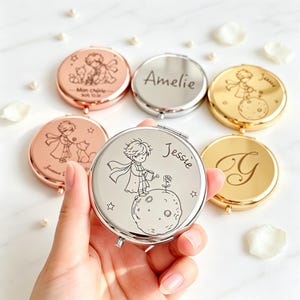 May include: A collection of round, metallic compact mirrors in rose gold, silver, and gold finishes. Each mirror features laser-engraved illustrations of a small prince character, stars, and roses, with some mirrors personalised with names like Amelie and Jessie.