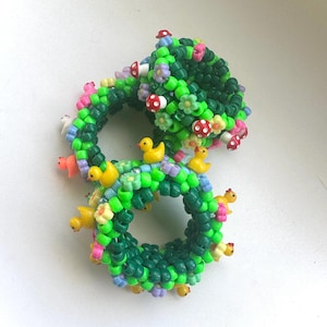 Puede incluir: Dos pulseras circulares hechas de cuentas de plástico verde vibrante. Las pulseras están decoradas con pequeños dijes de patitos amarillos, diminutas cuentas de hongos rojos y blancos, y flores de plástico de color violeta claro y amarillo.