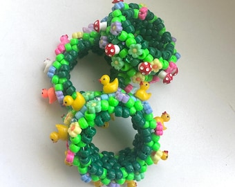 Può includere: Due braccialetti circolari realizzati con perline di plastica verde brillante. I braccialetti sono decorati con piccoli ciondoli a forma di papera gialla, minuscole perline a forma di fungo rosso e bianco e fiori di plastica lilla e gialli.