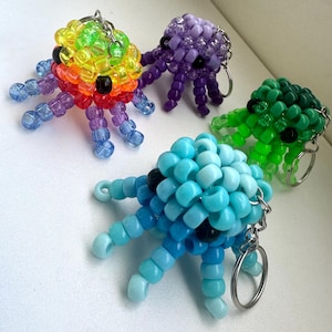 May include: Four octopus-shaped keychains crafted from colorful plastic pony beads. Each creature features a rounded head and eight dangling tentacles, with black beads for eyes. The set includes one rainbow, one purple, one green, and one blue octopus.