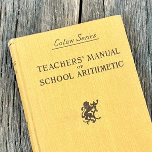 May include: A vintage, mustard-yellow book titled "TEACHERS' MANUAL of SCHOOL ARITHMETIC" from the Colaw Series. The cover features a small emblem and the publisher's name, B.F. Johnson Publishing Co., Richmond, Va. The book rests on a weathered, gray wooden surface.
