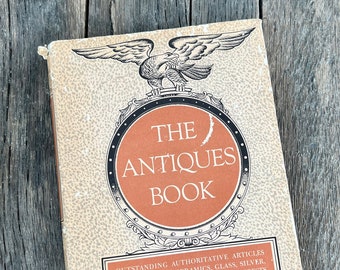 May include: A vintage book titled "The Antiques Book" with an illustration of an eagle on the cover. The book features articles on furniture, ceramics, glass, silver, pewter, and other collecting interests. It is edited by Alice Winchester and the staff of The Magazine Antiques.