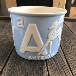 May include: A blue ceramic mug with a white letter A and the word "ANTELOPE" printed on the side. The letter A is embossed and the mug features a white silhouette of an antelope.