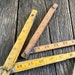 May include: Three vintage wooden folding rulers with yellow painted markings. The rulers are numbered from 1 to 28. The rulers are lying on a weathered wooden surface.