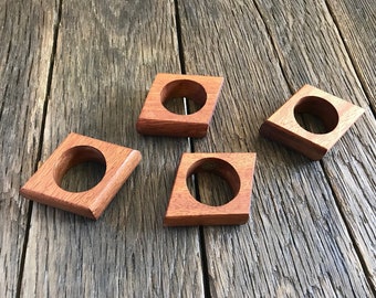 May include: Four wooden napkin rings in the shape of a diamond. The rings are made of a reddish-brown wood and have a smooth finish.
