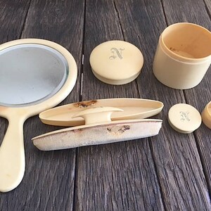 May include: A vintage vanity set with a hand mirror, a round compact, a small round container, a rectangular container with a brush, and a small round container with a lid. The set is made of ivory-colored plastic and has a delicate design.