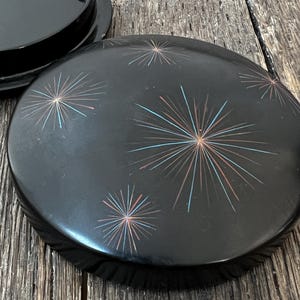 May include: A round black container lid featuring a decorative pattern of starbursts in light blue and copper tones. The lid is smooth and circular, with a matching black base component visible in the upper left corner of the frame.
