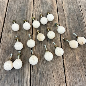 May include: A collection of fifteen round cabinet knobs featuring a white ceramic finish with a subtle crackle pattern. Each knob is attached to a short metal bolt with a brass-toned washer and nut assembly, providing a uniform look for furniture.