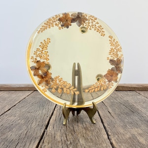 May include: A round, clear resin decorative piece with dried flowers and foliage embedded within. The piece is displayed on a small, gold-colored metal stand, showcasing the botanical design. The resin is a light yellow color, and the flowers are brown.