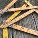 May include: Four wooden folding rulers with yellow painted markings. The rulers are numbered from 7 to 22. The rulers are lying on a weathered wooden surface.