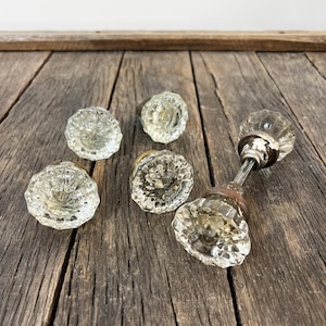 May include: A collection of five clear glass door knobs with a faceted, starburst-like pattern. Four of the knobs are individual pieces, while one knob is attached to a metal spindle and a circular metal base plate, showing the hardware assembly.