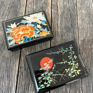May include: Two rectangular black lacquer boxes with glossy finishes. One box features a vibrant orange and white floral pattern with blue accents. The second box displays a red sun motif alongside green and yellow botanical illustrations.