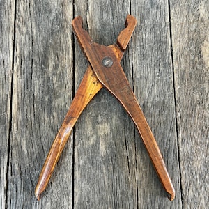 May include: A vintage wooden tool with a scissor-like design, featuring curved handles and a central pivot point. The tool is made of dark brown wood with a small metal rivet. The tool is set against a weathered wooden background.