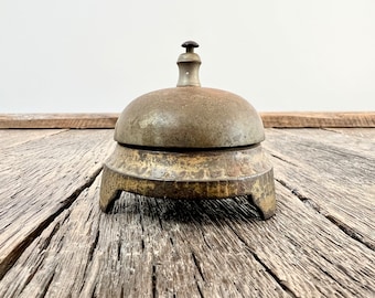 May include: An antique brass bell with a dome-shaped top and a small handle. The bell sits on a three-legged base. The bell has a weathered appearance, suggesting age and use. The bell is displayed on a rustic wooden surface.