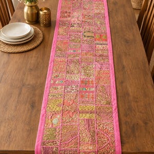 May include: A rectangular table runner featuring a patchwork design of various embroidered fabric squares in shades of pink, gold, and green. The runner is bordered by a solid pink fabric trim and displays intricate metallic thread detailing.