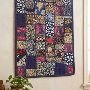 Op de afbeelding: Een rechthoekige wandhanger met een patchwork van diverse stoffen vierkanten in marineblauw, goud, roze en wit. Het textiel toont ingewikkeld borduurwerk, bloemmotieven en paisley-patronen, allemaal omrand door een effen marineblauwe bies.