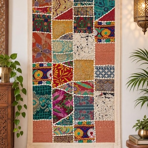 Könnte beinhalten: Ein rechteckiger Wandbehang mit einem Patchwork-Design aus verschiedenen bunten Stoffquadraten. Das Textil zeigt aufwendige Stickereien, florale Muster und geometrische Formen in Rot-, Blau-, Grün- und Goldtönen, eingefasst in einen cremefarbenen Rahmen.