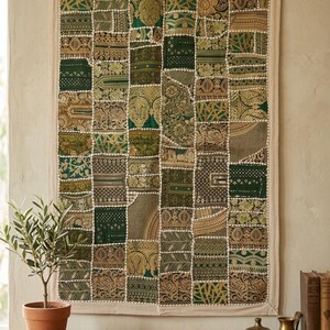 May include: A patchwork wall hanging with a beige border. The squares are various shades of green and gold with intricate patterns. The wall hanging is displayed on a light-colored wall. A small potted plant and decorative brass objects are on a wooden table.