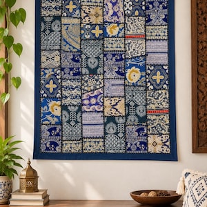 Op de afbeelding: Een rechthoekige wandhanger met een patchworkontwerp van diverse stoffen vierkanten in tinten diepblauw, goud en wit. Het textiel toont ingewikkelde bloem-, geometrische en paisley-patronen met zichtbare stiksels tussen elk vierkant.