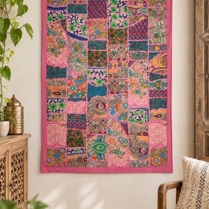 Puede incluir: Un tapiz rectangular con un diseño de retazos de varios cuadrados de tela bordados en tonos rosa, azul verdoso, azul y dorado. El textil está bordeado por un marco de tela rosa sólido y muestra intrincados patrones florales y de cachemira.