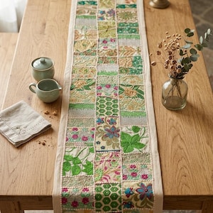 Può includere: Una tovaglia patchwork rettangolare con bordo beige e un design centrale di quadrati verdi e floreali. È posizionata su un tavolo di legno chiaro, con un piccolo vaso di fiori secchi e stoviglie in ceramica.