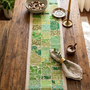 May include: A rustic wooden table is adorned with a green patchwork table runner. The runner features various shades of green in a square pattern. A bowl of decorative wooden beads, a small plate, a candle holder with a lit candle, and a napkin with a gold ring are on the table.
