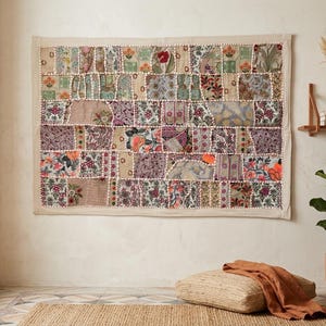 Puede incluir: Un gran tapiz de pared rectangular de patchwork. El textil presenta una variedad de patrones florales y geométricos en tonos beige, verde, morado y naranja. La pieza está enmarcada con un borde de color neutro.