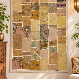 May include: A rectangular wall hanging featuring a patchwork design of various fabric squares in shades of gold, beige, and pink. The textile is embellished with intricate embroidery, small mirrors, beads, and white decorative trim borders.