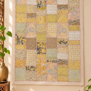 Puede incluir: Un tapiz rectangular con un diseño de patchwork de varios cuadrados de tela en tonos amarillo, crema y verde claro. Cada cuadrado muestra intrincados bordados, encajes o motivos florales, unidos por costuras visibles y pequeñas borlas.