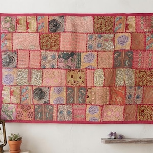 Pode incluir: Uma tapeçaria retangular com um design de patchwork de vários quadrados de tecido em tons de rosa, vermelho, dourado e castanho. O tecido apresenta padrões bordados intrincados, motivos florais e detalhes metálicos, tudo contornado por uma borda de tecido rosa sólido.
