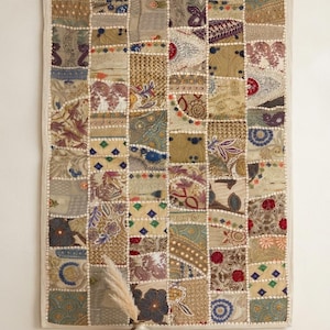 May include: A rectangular wall hanging featuring a patchwork design of various fabric squares in shades of gold, beige, blue, and red. The textile is composed of multiple patterned segments joined by white stitching, creating a grid-like appearance.