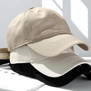May include: A stack of baseball caps in neutral colors: tan, off-white, and black. The caps are made of a smooth fabric and have a curved brim. The caps are stacked on top of each other, with the tan cap on top.