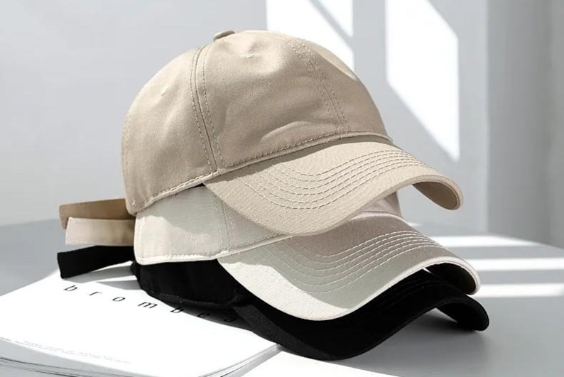 May include: A stack of baseball caps in neutral colors: tan, off-white, and black. The caps are made of a smooth fabric and have a curved brim. The caps are stacked on top of each other, with the tan cap on top.