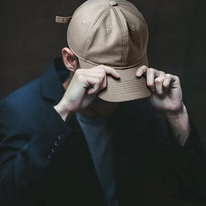 May include: A tan baseball cap with a curved brim. The cap is being held by a person wearing a dark blue blazer. The background is a dark, solid color.