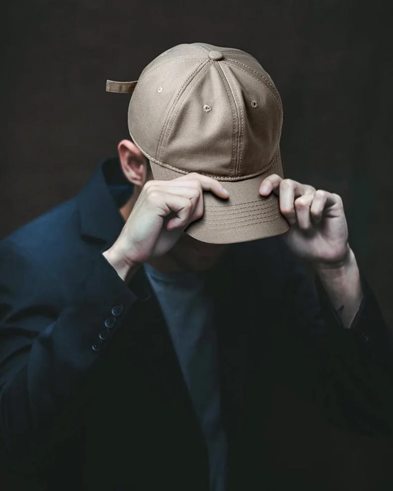 May include: A tan baseball cap with a curved brim. The cap is being held by a person wearing a dark blue blazer. The background is a dark, solid color.