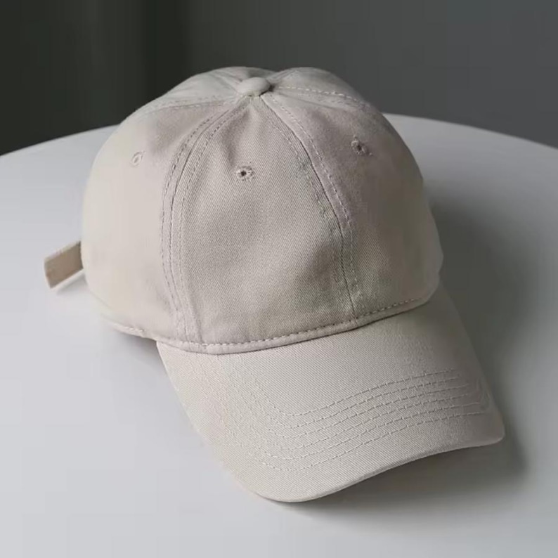 May include: A beige baseball cap with a curved brim. The cap is made of a soft fabric and features a matching adjustable strap at the back. The cap has a classic six-panel design with a button on top.