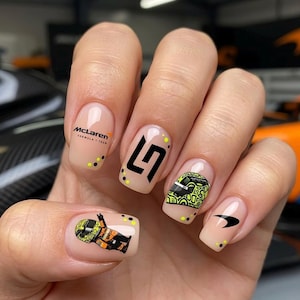 以下が含まれることがあります： ヌードカラーのネイルアート。McLaren Formula 1 Teamのロゴ、黒いLNロゴ、黄色と黒のレーシングヘルメットのグラフィック、レーシングスーツを着た小さなキャラクター、黒いマクラーレンのスピードマークロゴが描かれ、小さな黄色と黒のドットがアクセントになっています。