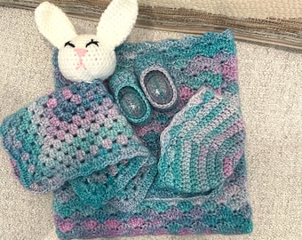 Può includere: Un set all'uncinetto composto da un doudou a forma di coniglio bianco con copertina, un paio di scarpine e un cappellino coordinati in tonalità sfumate di blu, verde e viola, realizzati con un motivo a punti testurizzati.