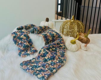 Può includere: Una sciarpa rettangolare all'uncinetto caratterizzata da un motivo in filato variegato nei toni del blu, crema e marrone. La sciarpa è esposta accanto a una collezione di zucche decorative, tra cui una zucca in filo dorato e diverse piccole zucche in tessuto bianco.