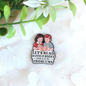 Peut inclure: Épingle en émail rectangulaire avec une illustration de dessin animé de deux enfants lisant un livre. L'épingle porte l'inscription "LET'S READ BANNED BOOKS AND CAUSE PROBLEMS" en noir et blanc. L'épingle est posée sur un fond floral blanc.