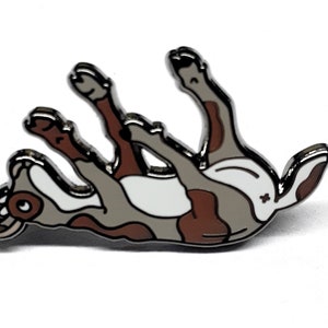 May include: A silver enamel pin of a brown and white dog with brown spots, lying on its back with its legs in the air.