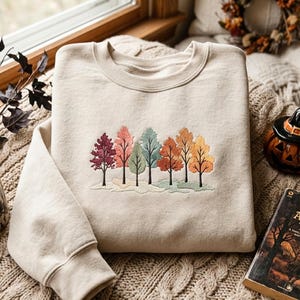 Könnte beinhalten: Ein beigefarbenes Sweatshirt mit Rundhalsausschnitt und einer Stickerei, die eine Reihe von sieben Bäumen in verschiedenen Farben wie Weinrot, Rosa, Türkis, Orange und Gelb zeigt, mit einer hellgrünen Bodenlinie unter den Stämmen.