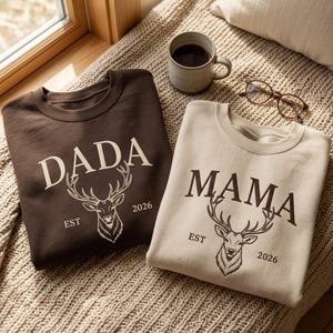 Peut inclure: Un sweat-shirt marron foncé avec le texte DADA et une illustration de cerf, et un sweat-shirt beige avec le texte MAMA et une illustration de cerf. Les deux portent le texte EST 2026. Une tasse en céramique et des lunettes sont posées à côté.