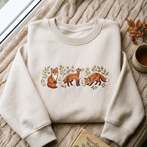 May include: A cream-colored crewneck sweatshirt featuring an embroidered design of three orange foxes surrounded by small green leaves, yellow flowers, and red berries. The sweatshirt has long sleeves with ribbed cuffs and a ribbed neckline.